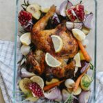 Overhead of tasty grilled whole chicken with lemon and pomegranate placed among onion with carrot served in glass bowl on towel